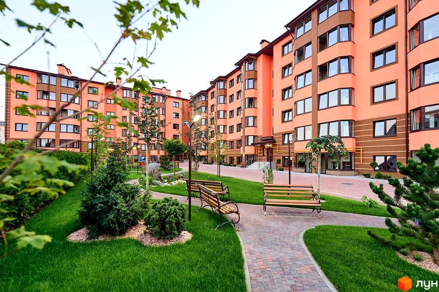 Новобудова Волошковий з фасадом персикового кольору та коричневими елементами. На прибудинковій території зелений газон, доріжки з бруківки та лавки для відпочинку.