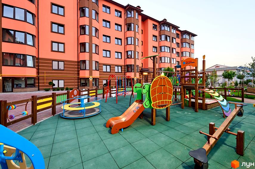 Житловий комплекс Волошковий: яскравий фасад будинку та сучасний дитячий майданчик з гірками, гойдалками та каруселлю. Зелене покриття та дерев'яні елементи створюють безпечний простір для дітей.