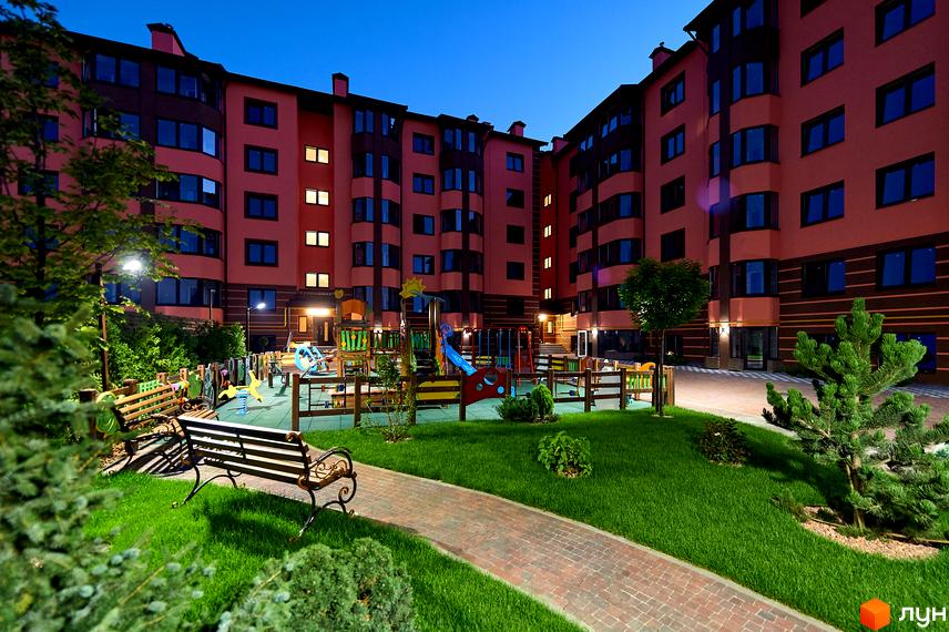 Житловий комплекс Волошковий. Фасад будинку в персикових тонах, вікна з темними рамами. Дитячий майданчик з гірками та гойдалками, лавки та зелені насадження на прибудинковій території.