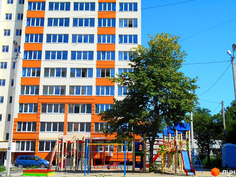 Житловий комплекс Затишна Оселя: багатоповерховий будинок з оранжевими та білими фасадами, дитячий майданчик та зелені насадження.