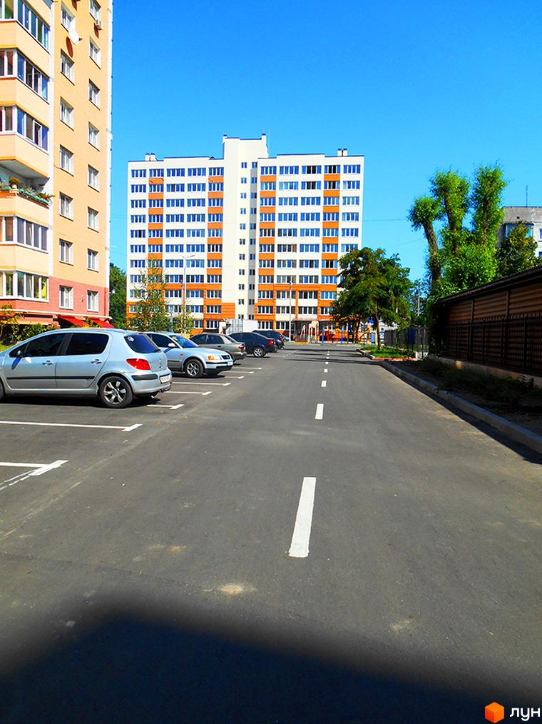 Зовнішній вигляд новобудови ЖК Затишна Оселя з парковкою та дорогою. Фотографія фасаду багатоповерхового будинку з вікнами та балконами.