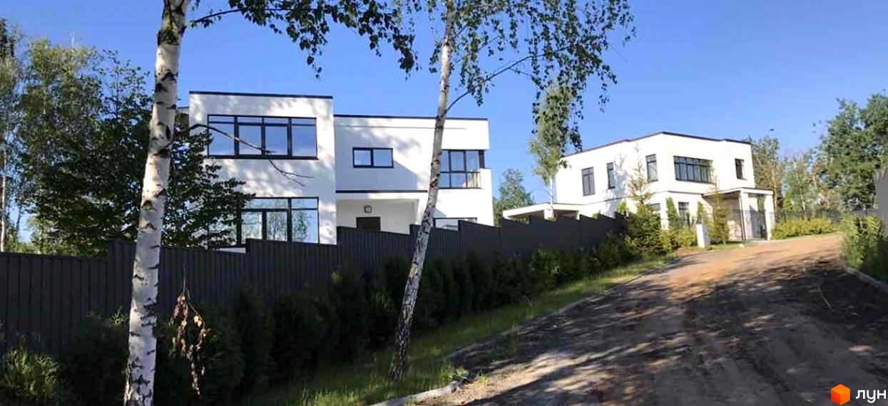 Білі двоповерхові будинки з темними вікнами та парканом у Котеджному містечку Barberry Park. Поруч ґрунтова дорога та дерева.