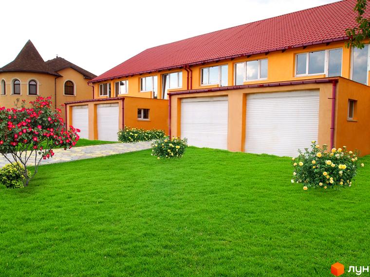 Зображення житлового комплексу по вулиці Комарова, 45. Двоповерховий будинок з гаражами та червоним дахом, перед будинком газон та декоративні кущі.