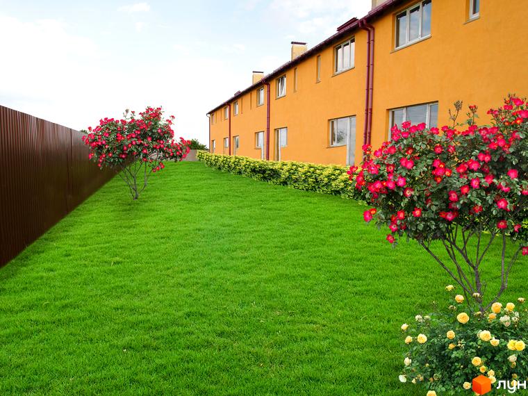 Вигляд будинку на Комарова, 45 з зеленим газоном та кущами троянд. Житловий комплекс з жовтими стінами та великими вікнами.