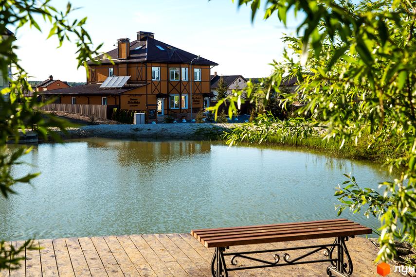 Котеджне містечко Home Resort: двоповерховий будинок з коричневим дахом та жовтими стінами, розташований біля озера з дерев'яним настилом та лавкою.