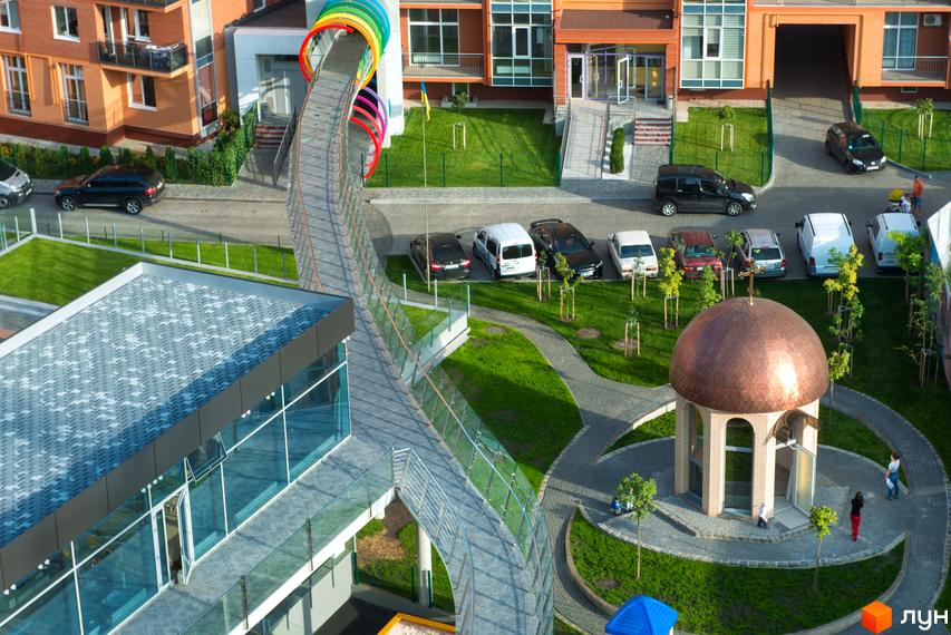 Житловий комплекс Водограй: багатоповерхові будинки з цегляними фасадами та скляною будівлею, прибудинкова територія з гіркою-тунелем, газонами та купольною спорудою.