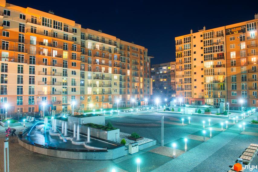 Житловий комплекс Водограй вночі. Сучасні багатоповерхові будинки з підсвіченими вікнами та балконами. На прибудинковій території розташований фонтан та ландшафтне освітлення.