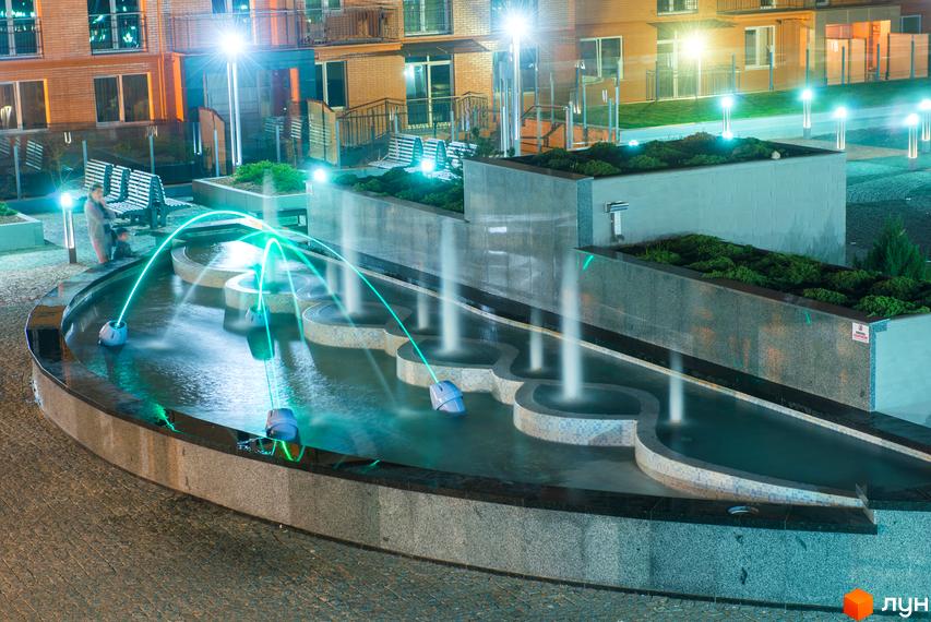 Житловий комплекс Водограй вночі з багатоповерховими будинками. На території розташований багаторівневий фонтан з зеленим підсвічуванням та ландшафтні зони.