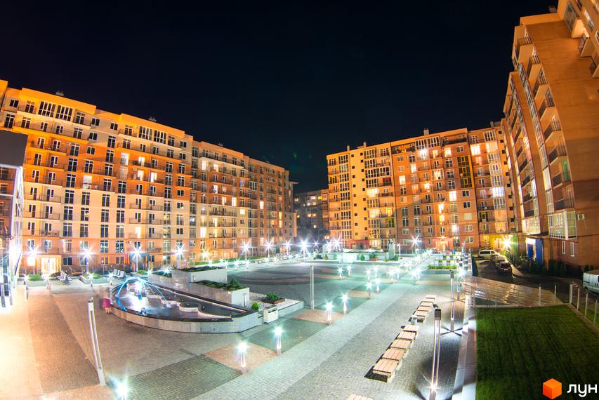Нічний вигляд житлового комплексу Водограй: багатоповерхові будинки світлих відтінків та освітлений внутрішній двір з бруківкою, ліхтарями та фонтаном.