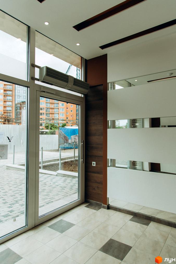 Вхідна група Житлового комплексу Водограй зі скляними дверима та видом на прибудинкову територію з бруківкою. На задньому плані видно багатоповерхові будинки.