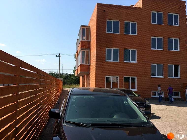 Зображення триповерхового цегляного будинку в Житловому комплексі Затишний Хотів. Перед будинком припарковані автомобілі та видно частину дерев'яного паркану.