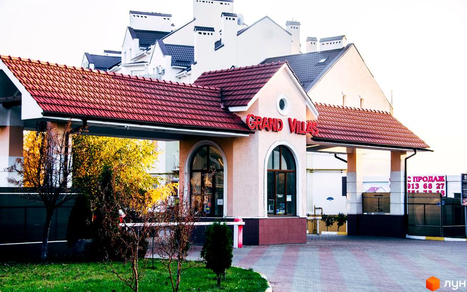Вхід до житлового комплексу Grand Villas з червоною покрівлею та прибудинковою територією. Зелені насадження та доріжки створюють комфортну атмосферу.