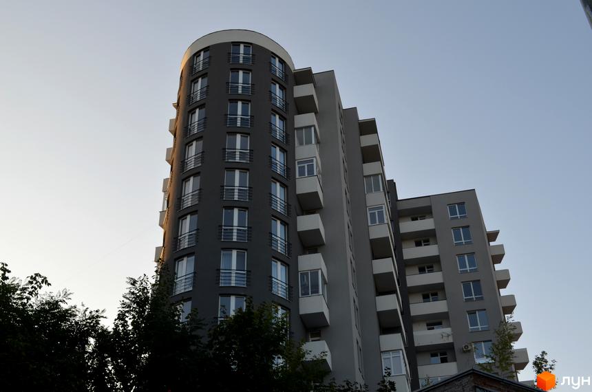 Зовнішній вигляд новобудови на вул. Водогінна, 2. Багатоповерховий будинок з балконами та вікнами на тлі неба. Навколо будинку зелені дерева.