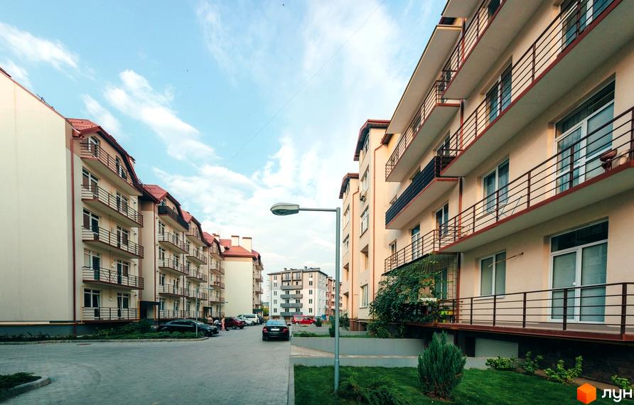 Житловий комплекс з балконами та терасами, збудований у світлих тонах. На прибудинковій території є газон, парковка та зелені насадження.