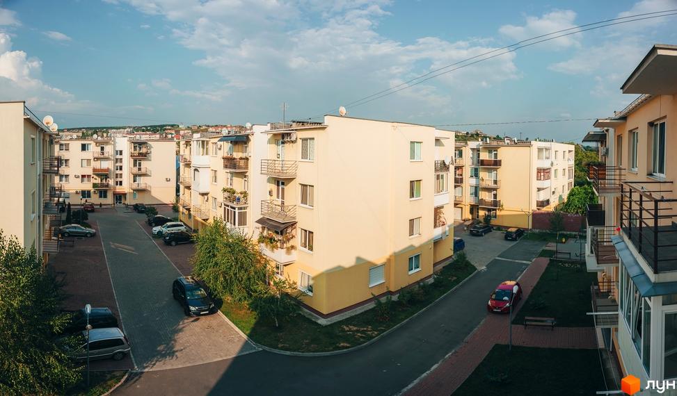 Вид на житловий комплекс з кількома багатоповерховими будинками з балконами та прибудинковою територією з асфальтованими доріжками та парковкою для автомобілів.