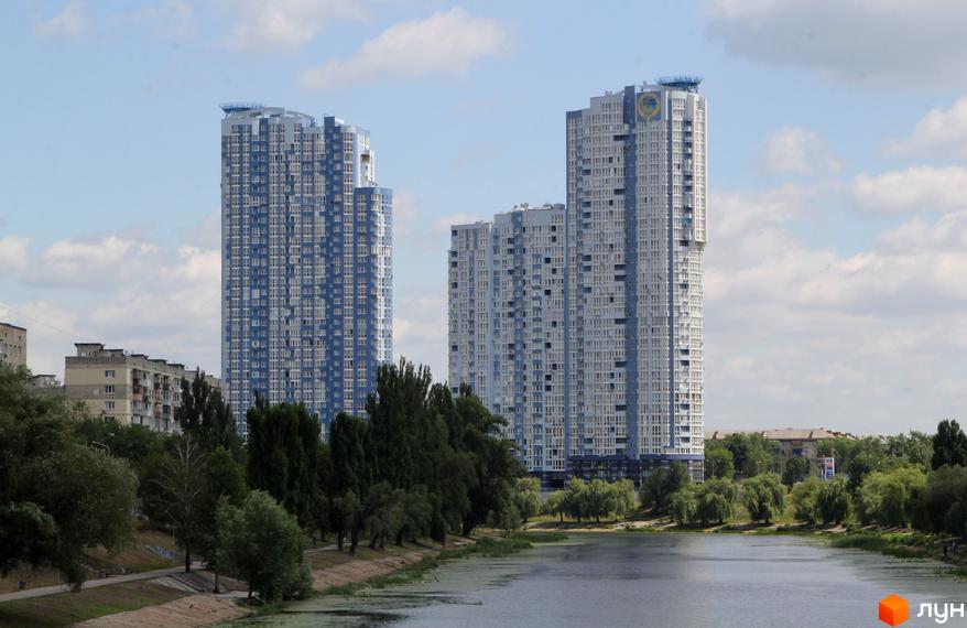 Київська Венеція - житловий комплекс з високими блакитними будівлями та великою кількістю вікон, розташований біля річки з зеленою зоною.