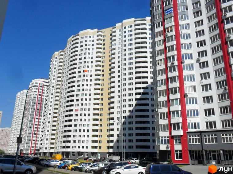 Житловий комплекс Позняки-4а: високі білі будівлі з червоними та сірими вставками, багато балконів та вікон. Навколо будівель розташована парковка з автомобілями.