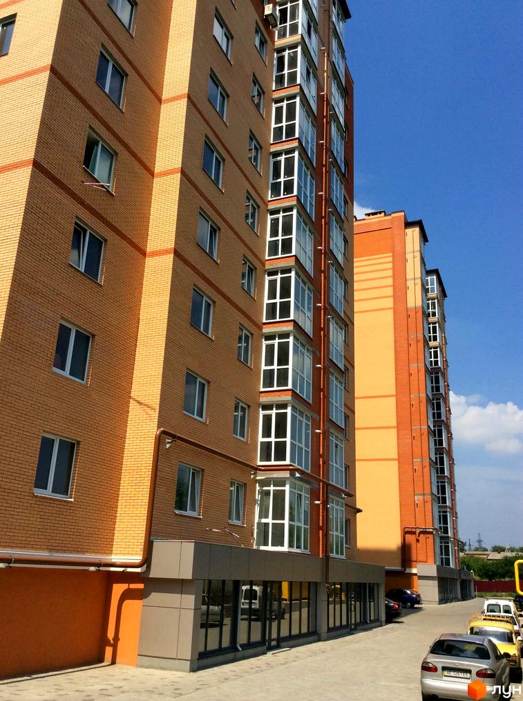 Висотна будівля ЖК Щасливий з цегляними фасадами та панорамним склінням. На прибудинковій території розташовані паркувальні місця для автомобілів.