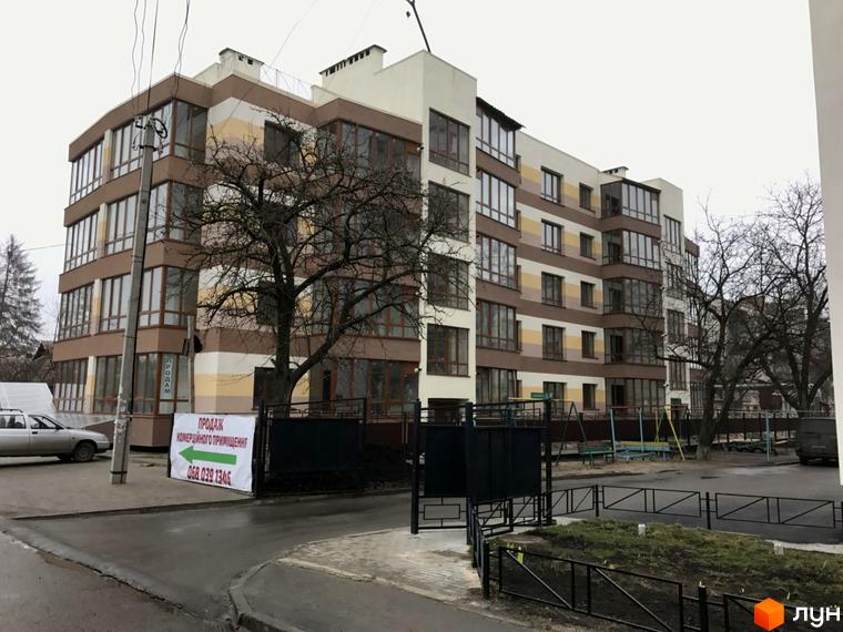 Багатоповерховий будинок з великими вікнами. На прибудинковій території є дитячий майданчик та парковка.