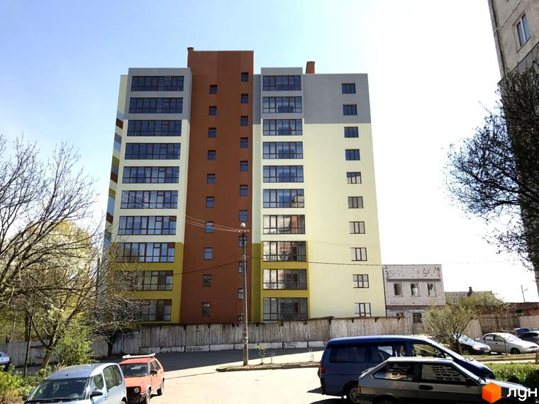 Житловий комплекс Holiday Park з фасадом, що поєднує коричневий, сірий та жовтий кольори. Навколо будинку розташовані дерева та припарковані автомобілі.