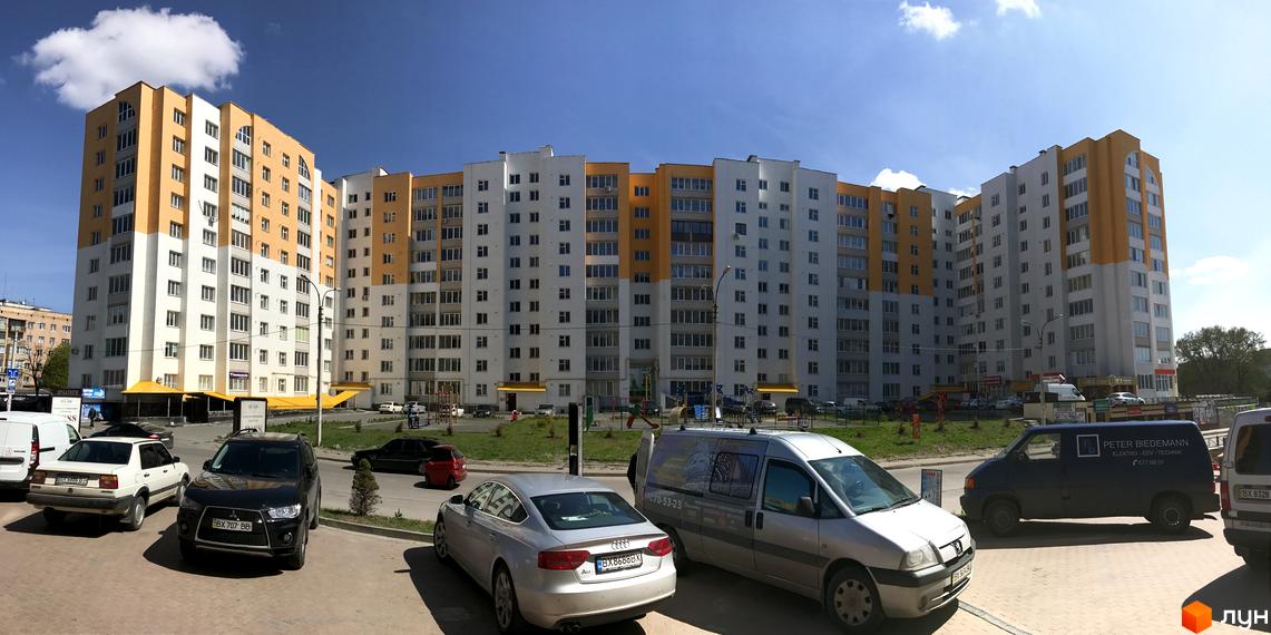 Зовнішній вигляд багатоповерхового житлового комплексу з білими та помаранчевими фасадами. На прибудинковій території розташовані автомобілі та дитячий майданчик.