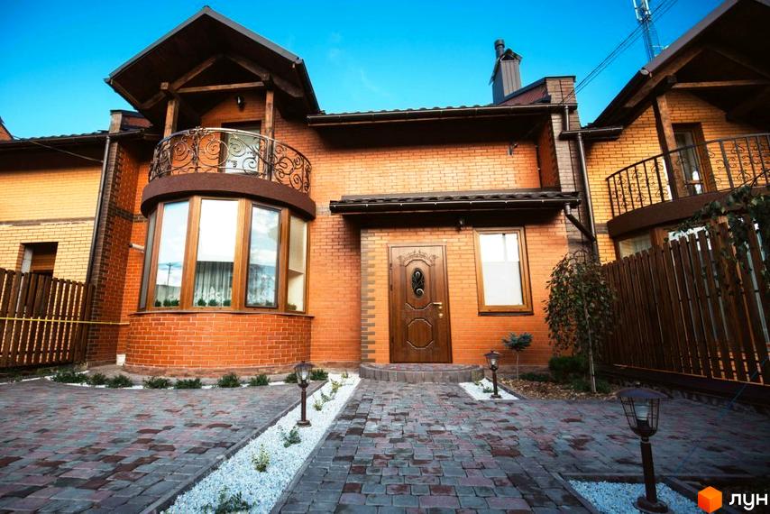 Котеджне містечко Sun House. Фасад будинку з червоної цегли з балконом та великими вікнами. Перед будинком вимощена бруківкою доріжка з ліхтарями та декоративними рослинами.