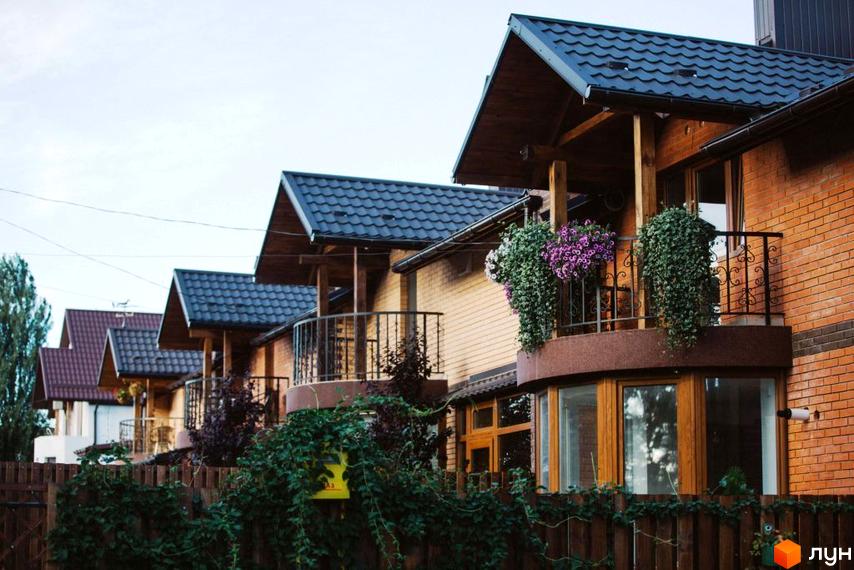 Котеджне містечко Sun House: фасади будинків з цегли та дерева, балкони з квітами та зеленню, дерев'яний паркан. Зовнішній вигляд новобудови.