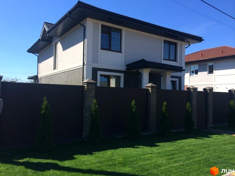 Двоповерховий будинок з білим та темно-сірим фасадом, темним дахом. Перед будинком зелений газон та коричневий паркан з кам'яними стовпами Котеджного містечка Петропавлівський Luxe.