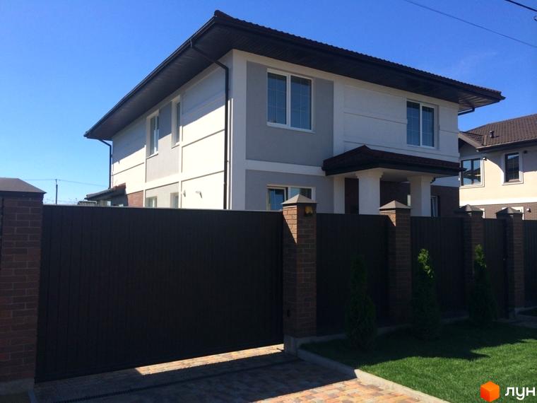 Двоповерховий будинок Котеджного містечка Петропавлівський Luxe з біло-сірим фасадом та темно-коричневим дахом. Перед будинком коричневий паркан, цегляні стовпи та зелений газон.