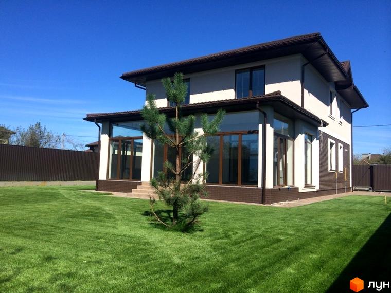 Двоповерховий будинок світлого кольору з коричневим дахом та великими вікнами у Котеджному комплексі Петропавлівський Luxe. Навколо будинку доглянутий зелений газон.