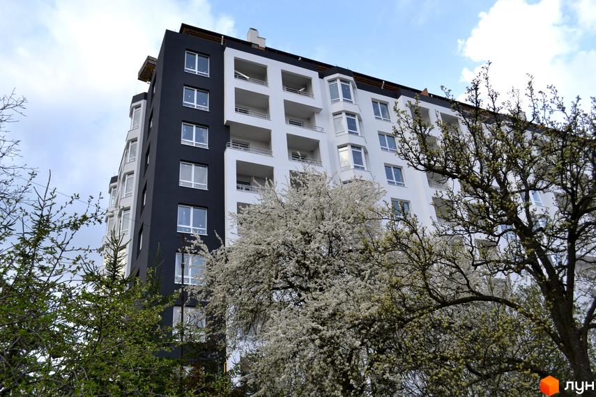 Житловий комплекс Kaiser - це сучасна новобудова з біло-чорним фасадом та балконами. Навколо комплексу розквітли дерева.