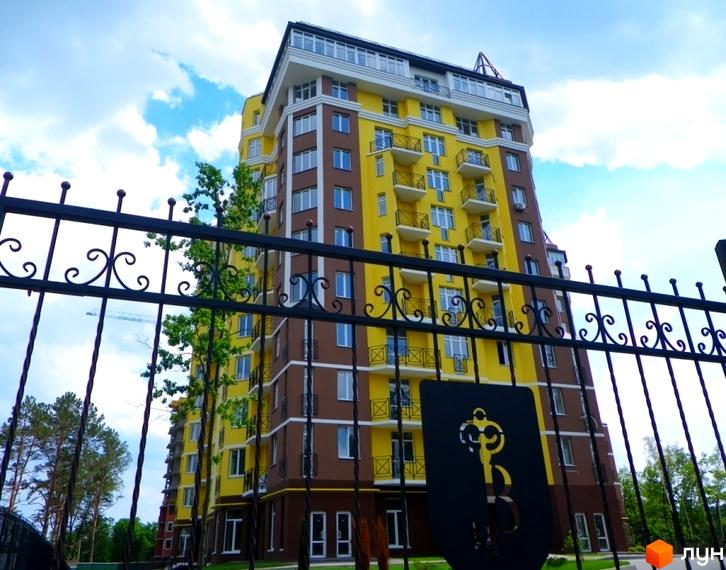 Житловий комплекс Велесгард: жовто-коричнева багатоповерхівка з балконами за чорною кованою огорожею. Навколо зелені дерева та блакитне небо з хмарами.