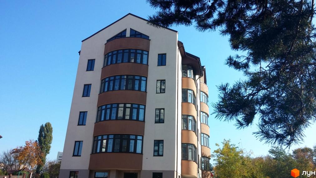 Фасад житлового комплексу Комфорт Сіті з вигнутими балконами та великими вікнами на тлі блакитного неба. Поруч ростуть дерева.