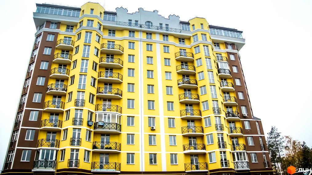 Фасад яскравого жовто-коричневого житлового комплексу Велесгард з численними балконами та великими вікнами. Будинок має незвичайну архітектуру з виступаючими елементами.