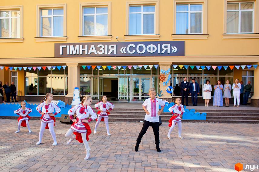 Житловий комплекс Софія Сіті: діти в національних костюмах танцюють біля жовтої будівлі гімназії. На фасаді будівлі вивіска «ГІМНАЗІЯ «СОФІЯ».