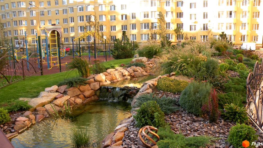 Житловий комплекс Софія Клубний. Фасад жовтої багатоповерхівки, дитячий майданчик та ландшафтний дизайн з водоймою та рослинністю.