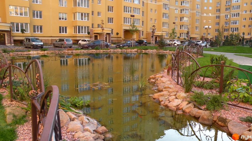 Житловий комплекс Софія Клубний з жовтими фасадами та озелененою прибудинковою територією з водоймою та декоративною огорожею.