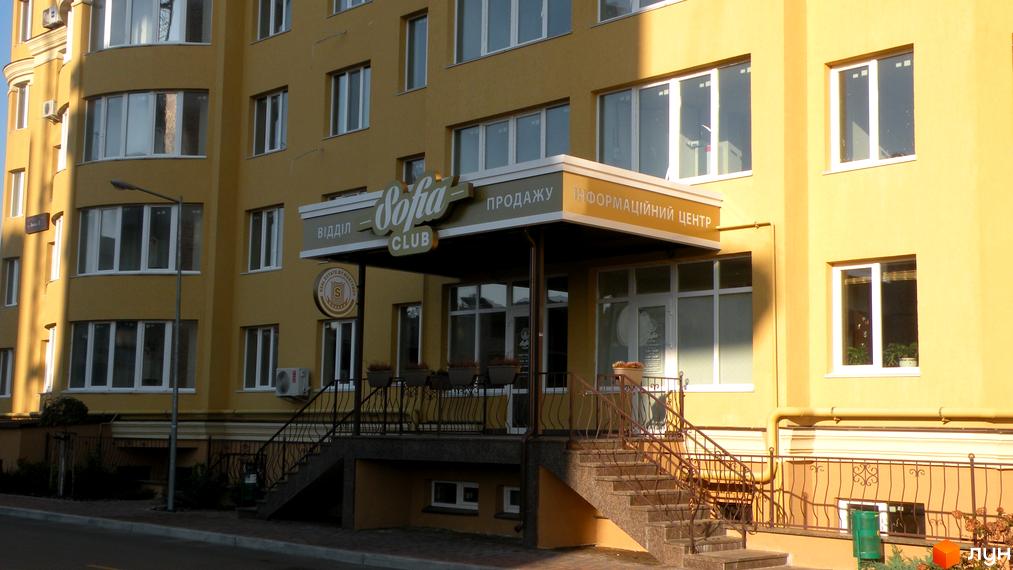 Житловий комплекс Софія Клубний: жовта фасадна частина будівлі з великими вікнами та вхідною групою зі сходами. Навколо будівлі розташовані зелені насадження та огорожа.