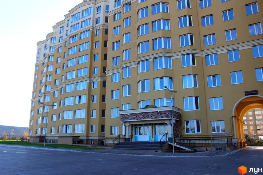 Зовнішній вигляд новобудови ЖК Софія Клубний з жовтим фасадом та численними вікнами. Прибудинкова територія з асфальтованим покриттям та зеленими насадженнями.