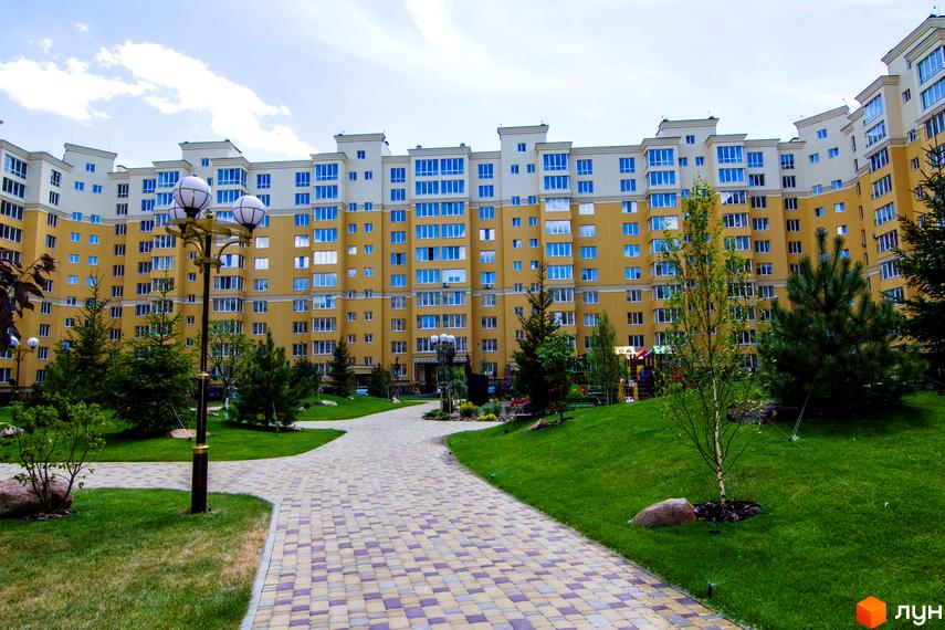 Житловий комплекс Софія Клубний. Фасад жовтої новобудови з блакитними вікнами та прибудинкова територія з доріжкою та зеленими насадженнями.