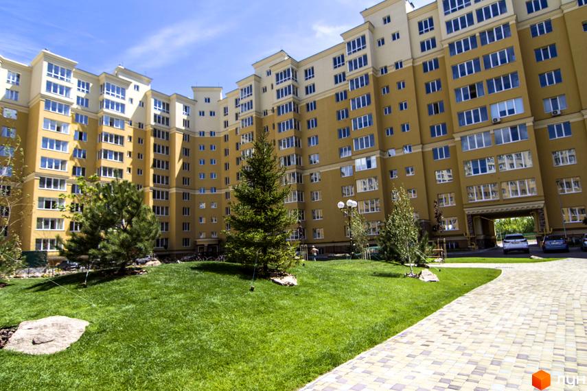 Житловий комплекс Софія Клубний: жовта багатоповерхівка з білими елементами та великою кількістю вікон. Доглянута прибудинкова територія з газоном, деревами та бруківкою.