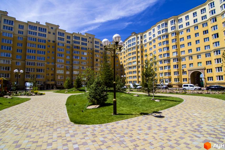 Житловий комплекс Софія Клубний. Зовнішній вигляд жовтої багатоповерхівки з синіми вікнами та прибудинкова територія з бруківкою та газоном.