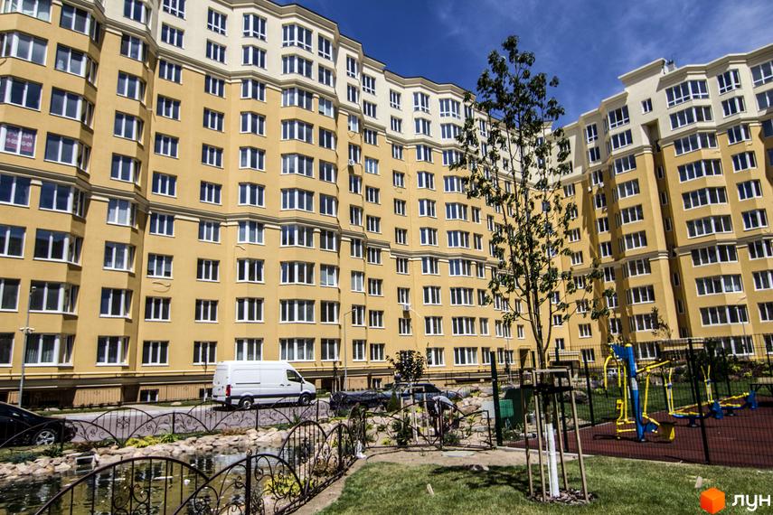 Житловий комплекс Софія Клубний: жовта багатоповерхівка з білими елементами та численними вікнами. Прибудинкова територія з деревами, газоном та спортивним майданчиком.