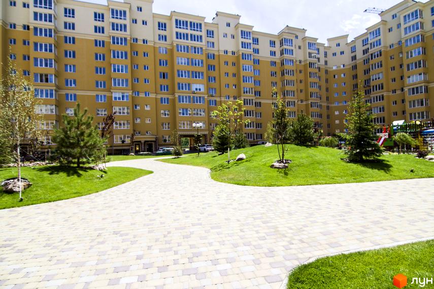 Житловий комплекс Софія Клубний з жовтим фасадом та блакитними вікнами. На прибудинковій території зелений газон, доріжки з бруківки та дитячий майданчик.