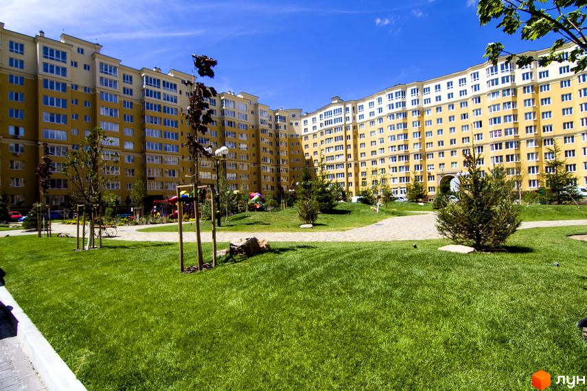 Житловий комплекс Софія Клубний. Зовнішній вигляд жовтої багатоповерхівки з численними вікнами та доглянутою зеленою прибудинковою територією з деревами та дитячим майданчиком.