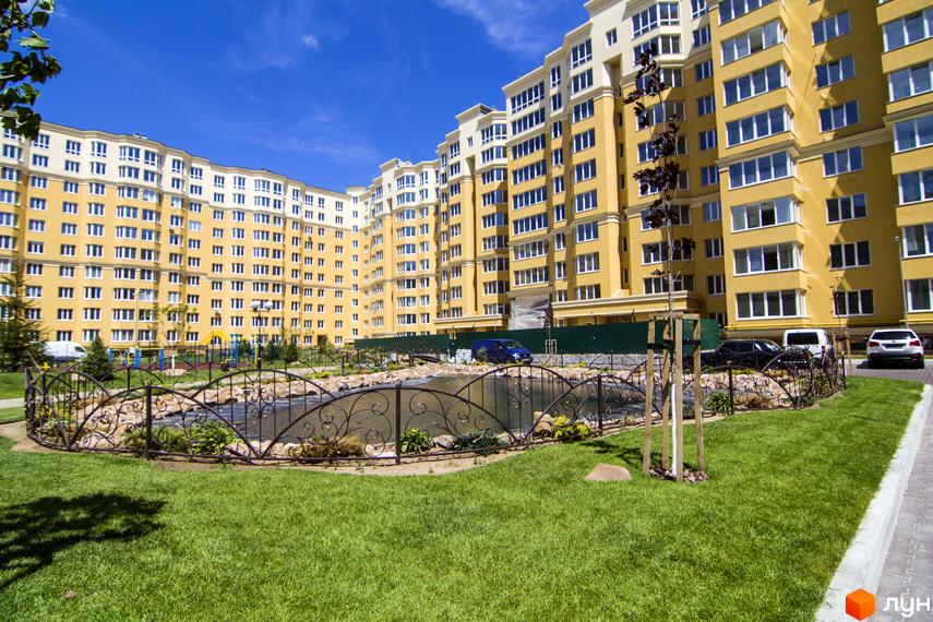 Житловий комплекс Софія Клубний. Фасад жовтого кольору з білими елементами, великі вікна. Доглянута прибудинкова територія з газоном, декоративним парканом та водоймою.