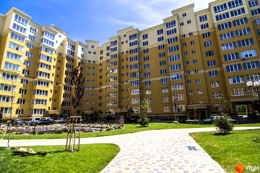 Зовнішній вигляд новобудови ЖК Софія Клубний з жовтими фасадами та великою кількістю вікон. Прибудинкова територія з доглянутим газоном та вимощеною доріжкою.