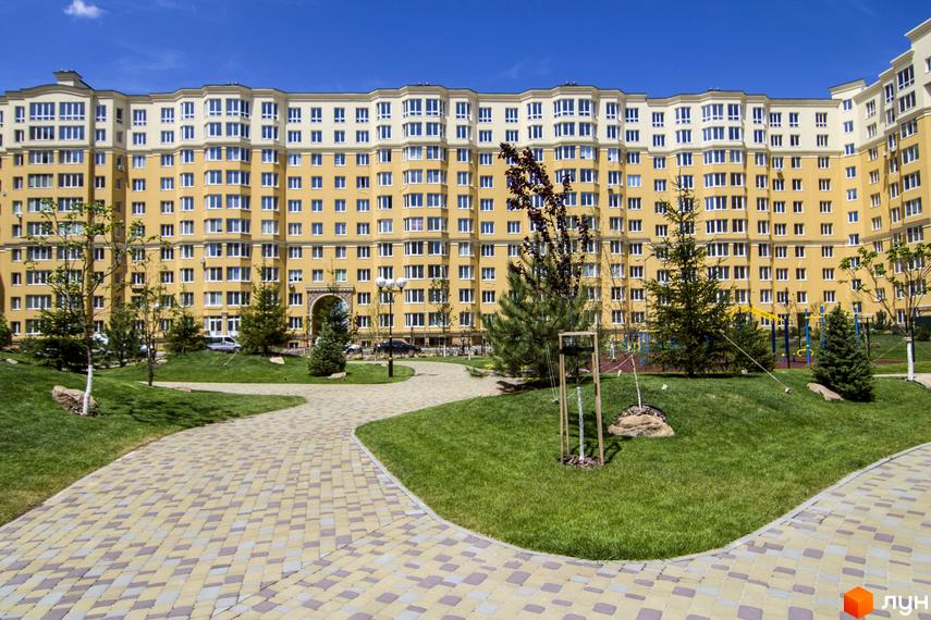 Зовнішній вигляд жовто-білої новобудови ЖК Софія Клубний з численними вікнами. На прибудинковій території розташовані зелені газони, доріжки з бруківки та дитячий майданчик.