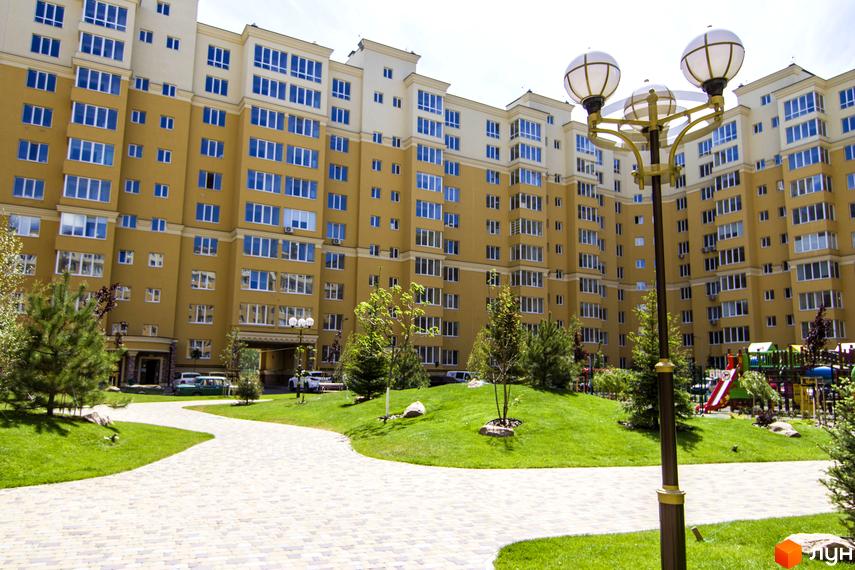 Житловий комплекс Софія Клубний. Зовнішній вигляд багатоповерхового будинку з жовтим фасадом та синіми вікнами. Прибудинкова територія з зеленим газоном, доріжками та дитячим майданчиком.