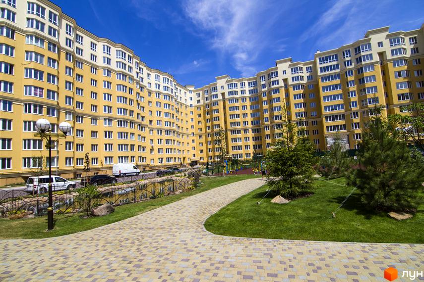 Житловий комплекс Софія Клубний з жовтим фасадом та білими елементами, що має багато вікон. Прибудинкова територія з доглянутим газоном, доріжками та деревами.